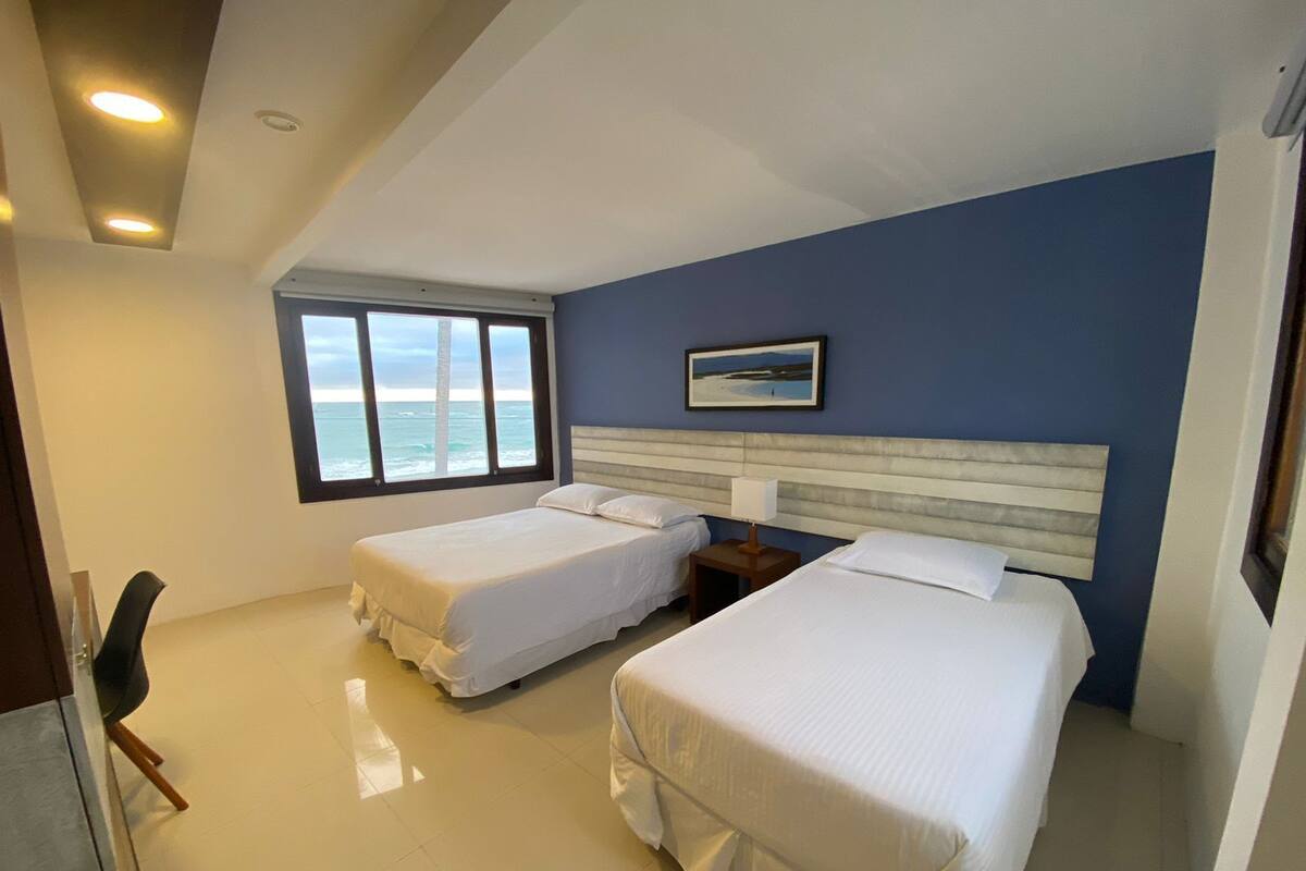 A hotel room with two beds, a large window with a view of the sea, a painting on the wall, and a small bedside table with a lamp.