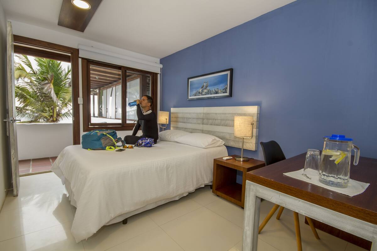 A hotel room with a bed, a nightstand, a table, a window with a view of palm trees, and a painting on the wall.