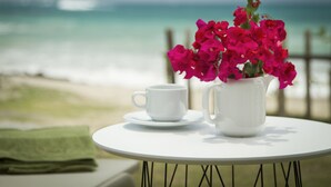 Ein weißer Tisch mit einer weißen Tasse und Untertasse sowie einer weißen Kanne mit rosa Blumen, vor einer Strandkulisse.