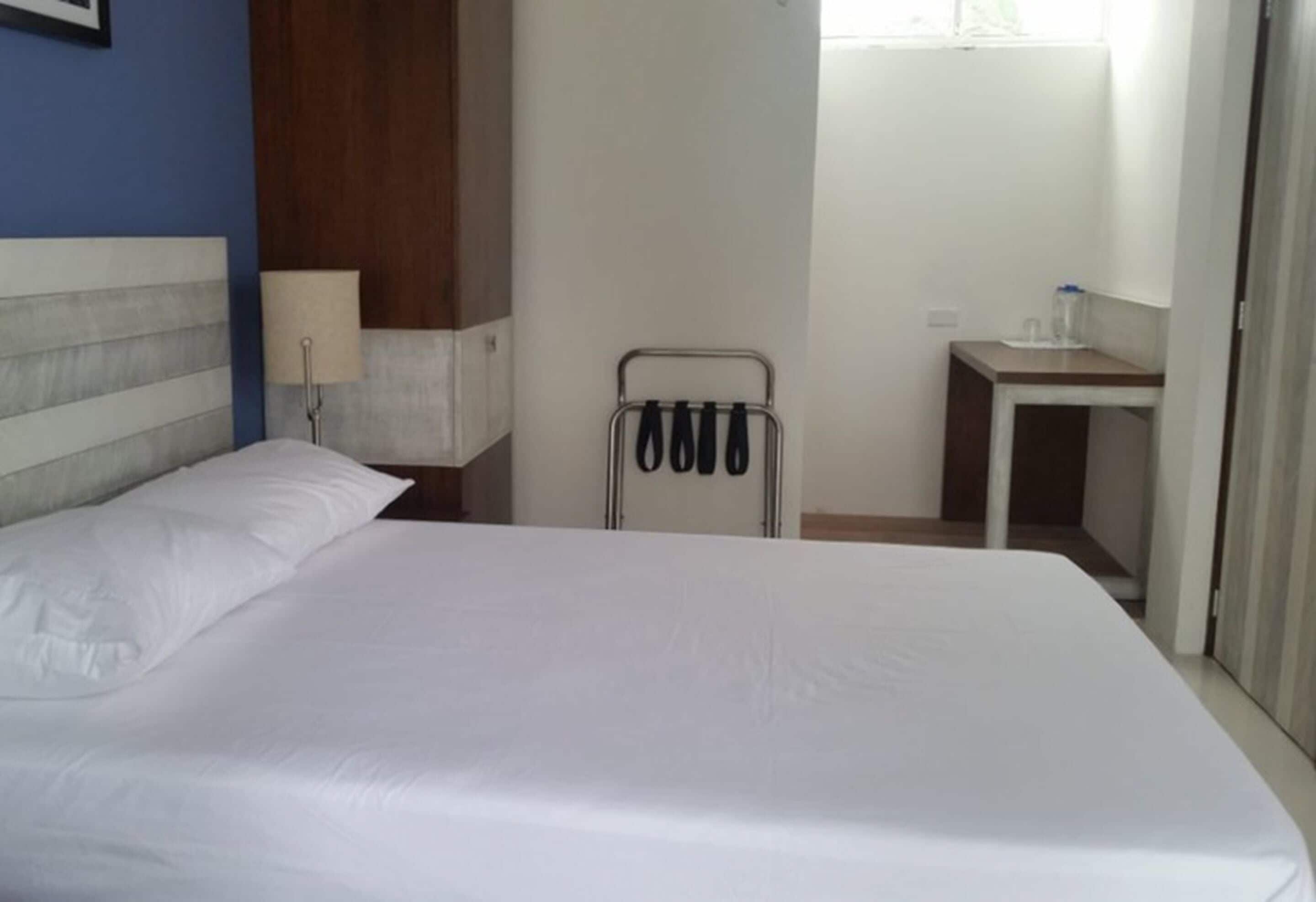 Una habitación de hotel con una cama grande, una mesita de noche, una lámpara, un perchero para toallas y un pequeño baño.