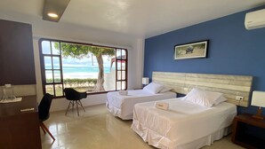 Ein Hotelzimmer mit zwei Betten, einem Schreibtisch und einem Sessel. Ein großes Fenster bietet Blick auf den Strand und Palmen.