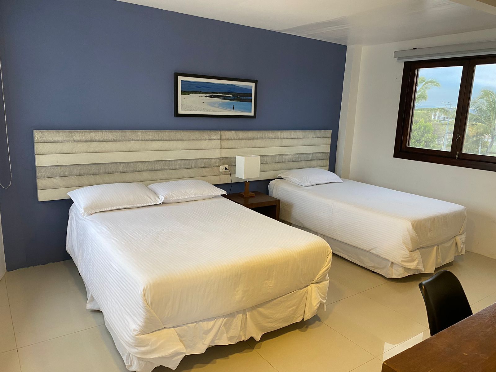 A hotel room with two beds, a headboard, a nightstand, a lamp, and a window with a view of palm trees.