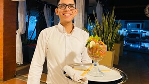 A person in a white shirt holding a tray with a dessert and a drink, in a room with a tiled floor and a plant in the background. - Farallones Hotel (Chinandega)