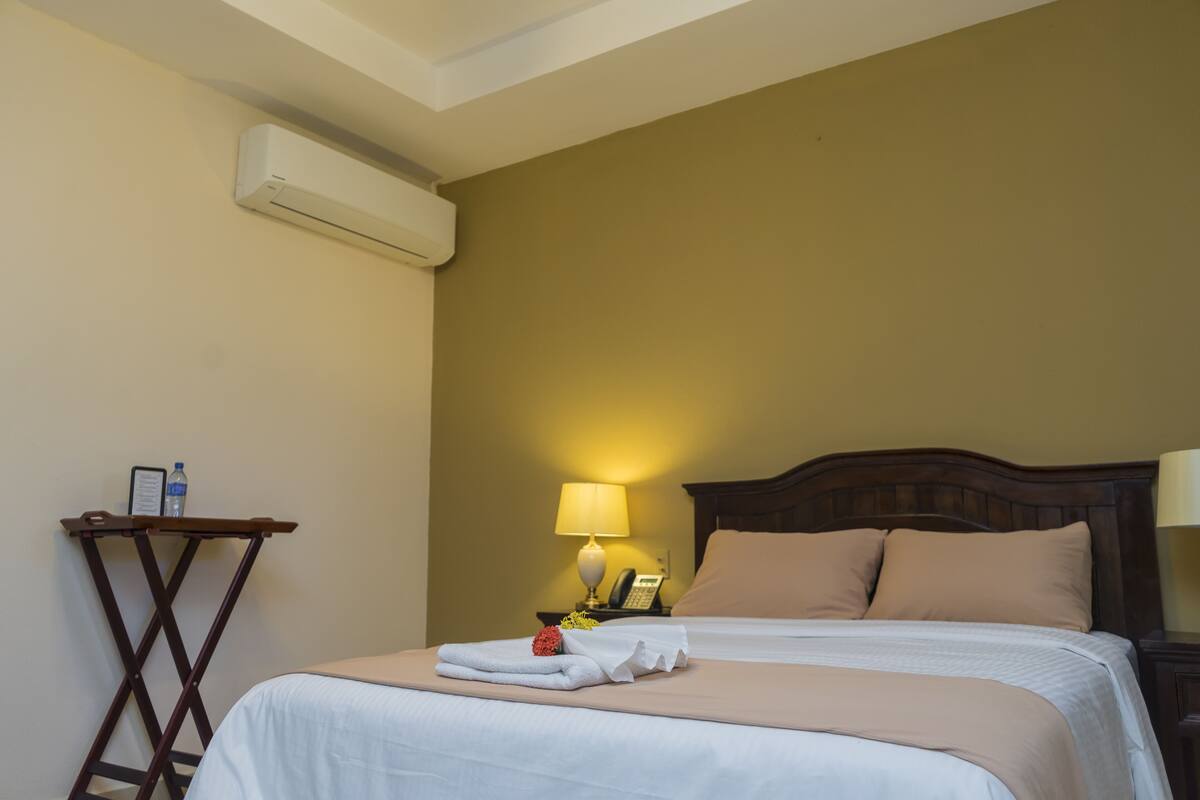 A hotel room with a bed, a nightstand with a lamp, a telephone, and an air conditioning unit on the wall.