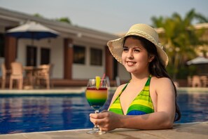A person sitting by a pool with a drink, wearing a striped swimsuit and a straw hat. - Farallones Hotel (Chinandega)