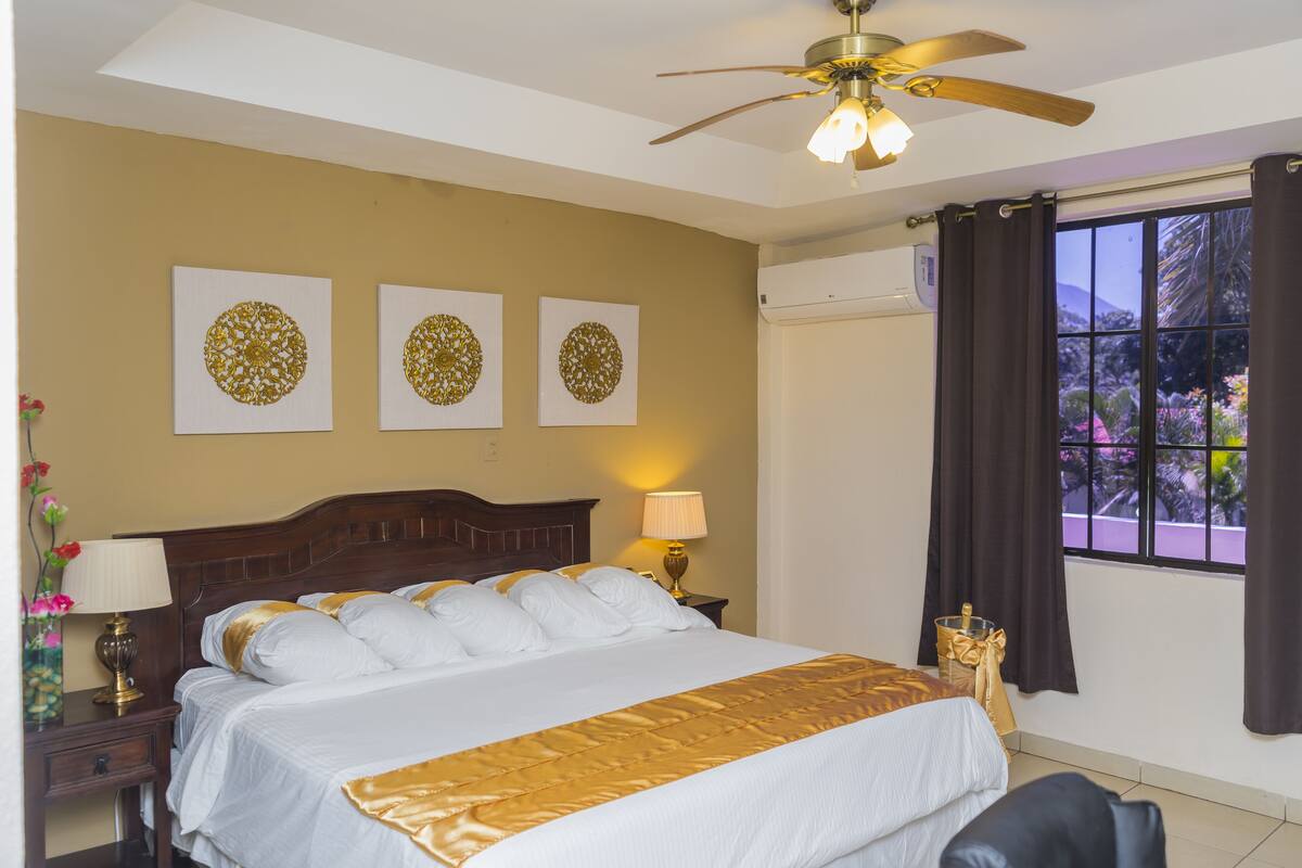 A hotel room with a bed, a ceiling fan, a window with curtains, and wall art.