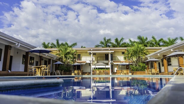 A pool area with lounge chairs and umbrellas, surrounded by palm trees and a building with balconies. - Farallones Hotel (Chinandega)