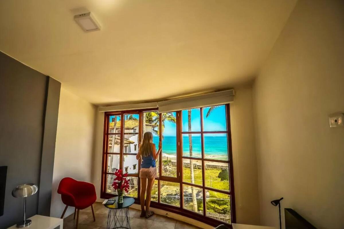 A room with a large window offering a view of the beach and palm trees.