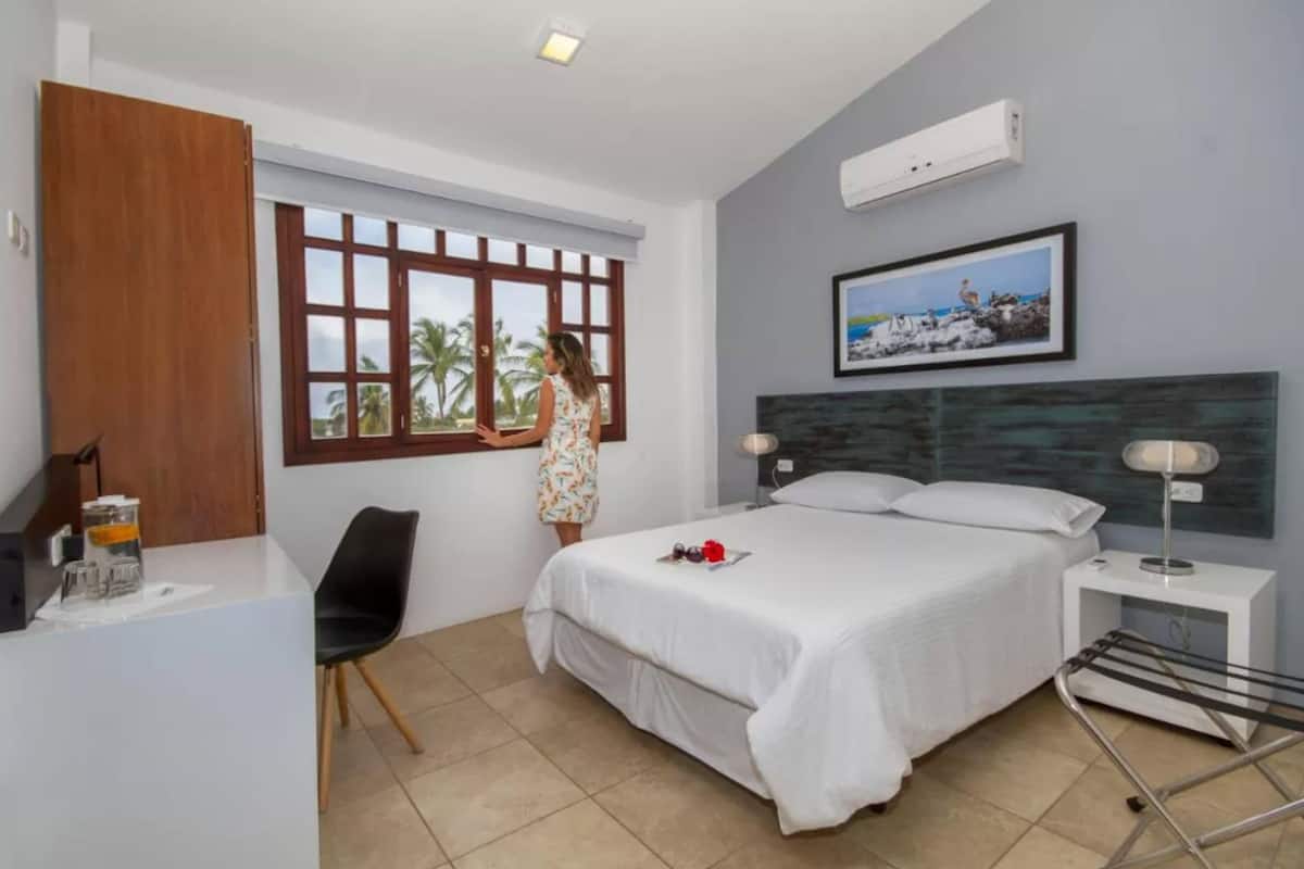 A hotel room with a bed, a desk, a chair, a window with a view of palm trees, and a painting on the wall.