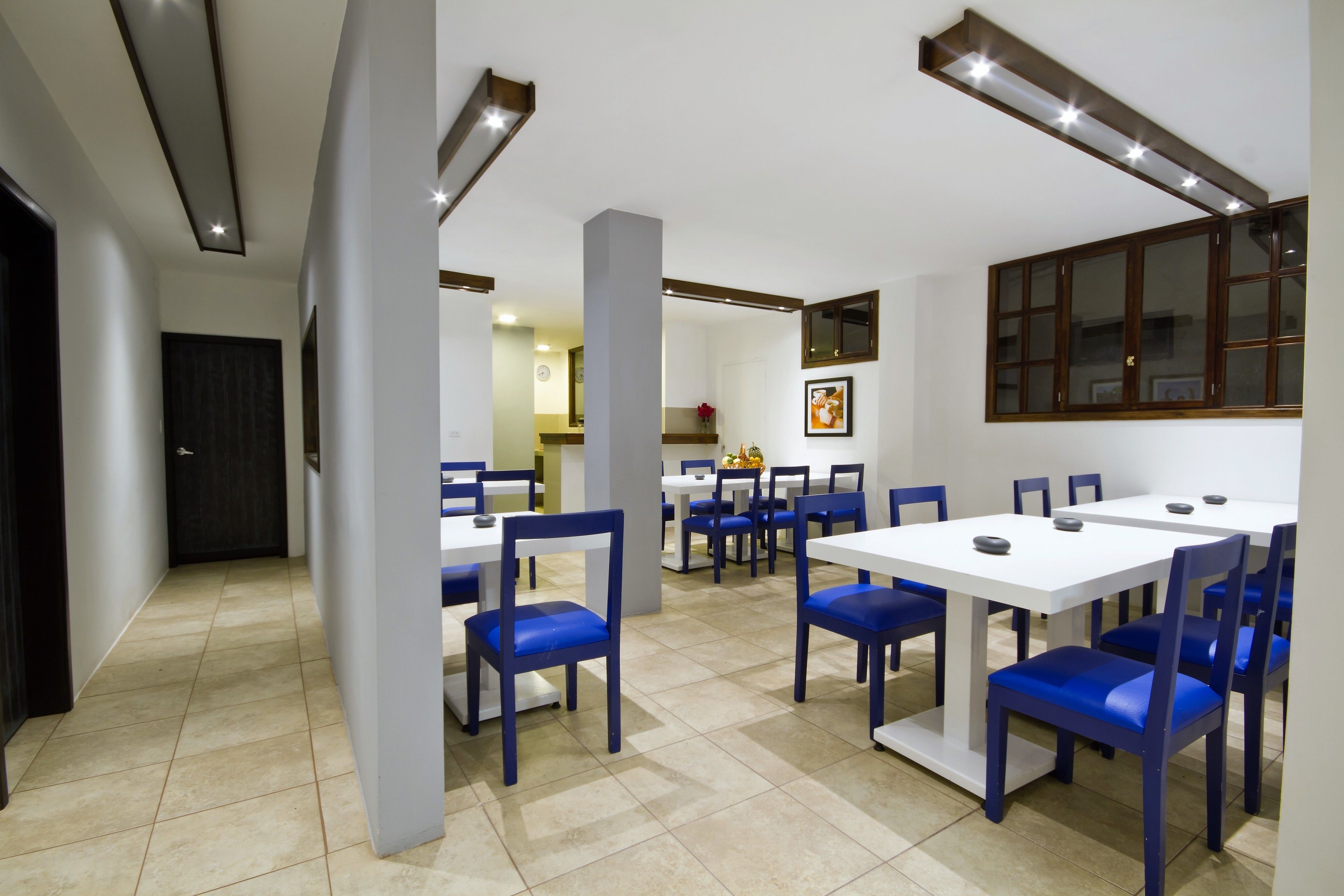 A dining area with white tables and blue chairs, a kitchen in the background, and a large window with blinds.