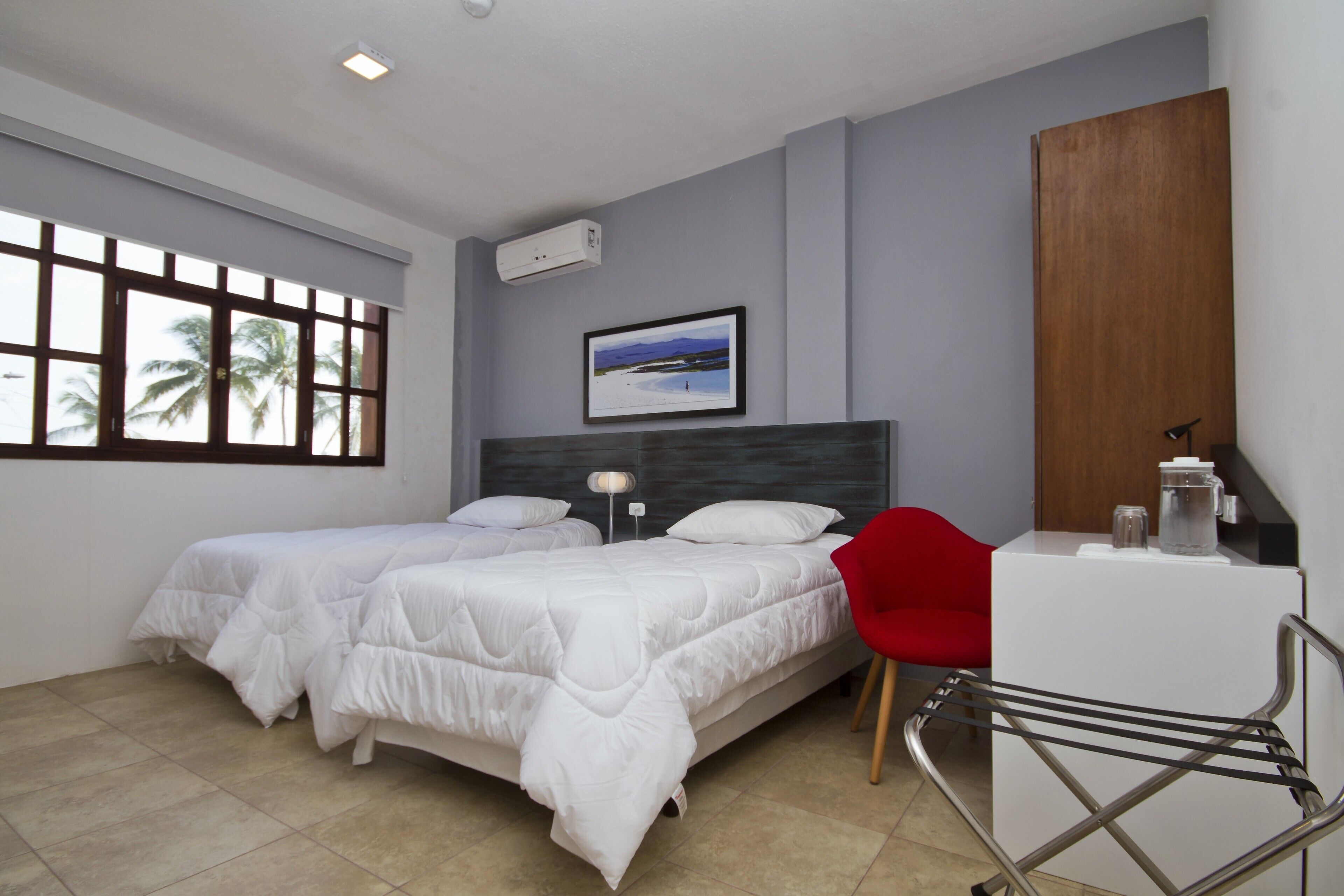 Habitación de hotel con dos camas, un escritorio, una silla, una ventana con vistas a palmeras y un cuadro en la pared.