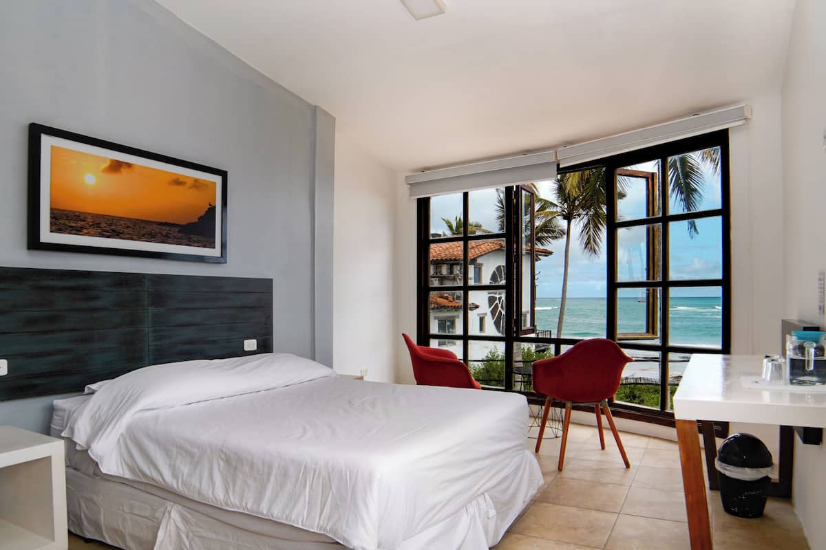 A hotel room with a bed, a desk, a chair, and a large window with a view of the ocean.