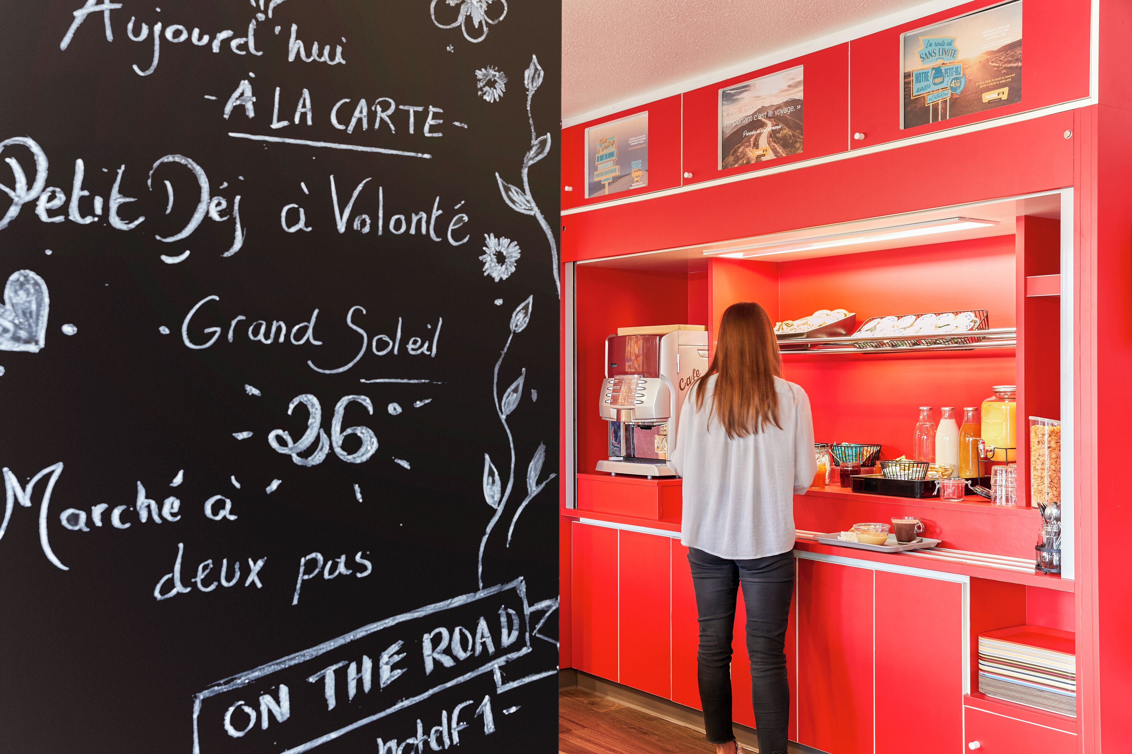 Un intérieur aux couleurs rouge et noir, avec un tableau noir sur lequel est écrit du texte, et une personne debout derrière un comptoir, devant lequel se trouvent divers objets.