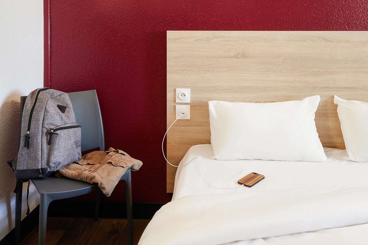 A hotel room with a wooden headboard, a bed with white bedding, a gray backpack on a chair, and a phone on the bed.