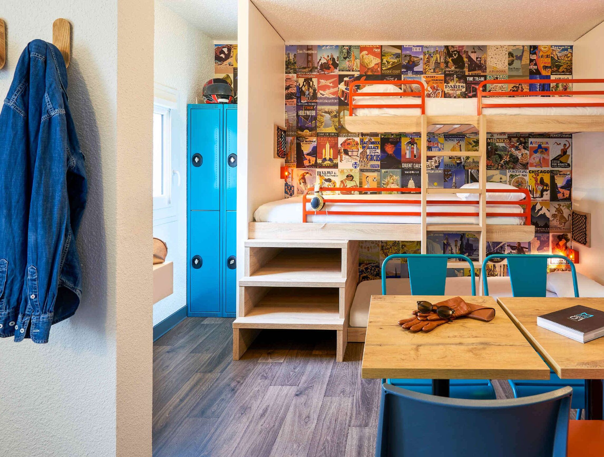 Une petite pièce avec des lits superposés, un bureau et une chaise. On y trouve un placard bleu et un mur orné d’affiches de voyage.
