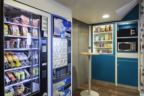 A kitchenette with a microwave, shelves stocked with snacks, and a vending machine with beverages and snacks. - hotelF1 Nantes EST La Beaujoire  (Nantes)