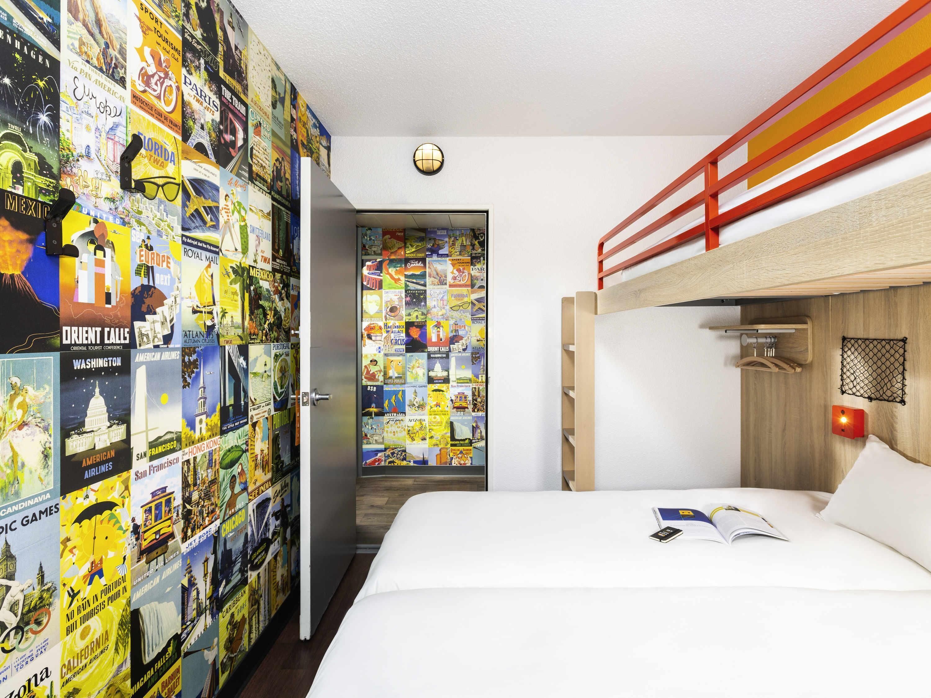 Une chambre avec un lit superposé, un mur recouvert d’affiches de voyage, une porte avec un miroir et une table de chevet sur laquelle se trouvent un livre et un téléphone.