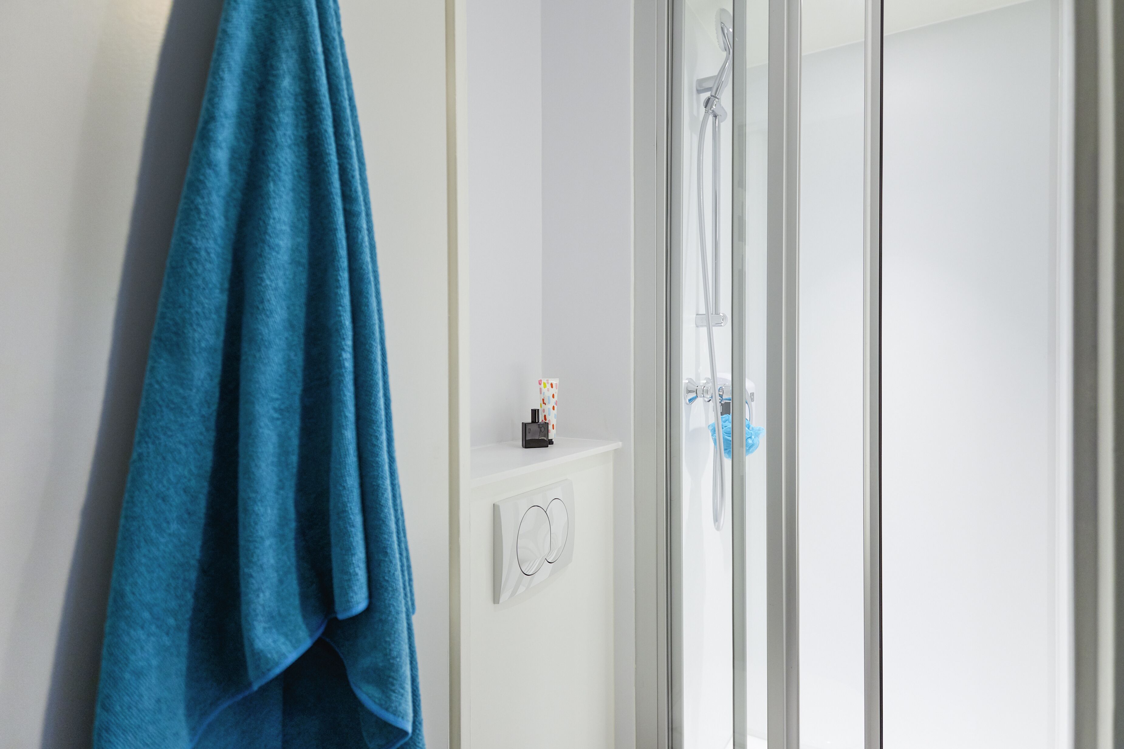 A bathroom with a glass shower enclosure, a towel hanging on the wall, and a small shelf with toiletries.