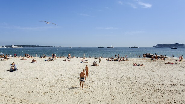 Een strand met mensen die zonnebaden en wandelen, boten in het water, en een heldere lucht.