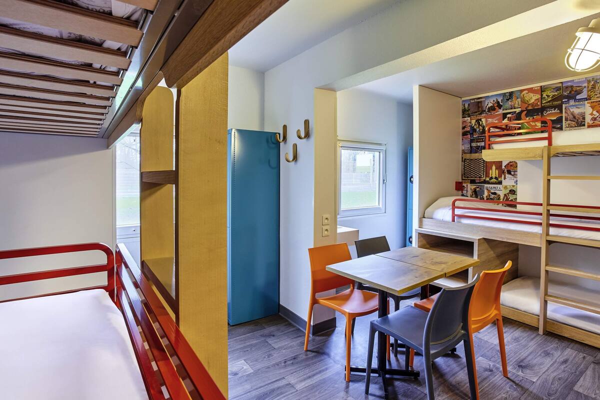 A compact room with bunk beds, a small table, and chairs. There is a window and a door.