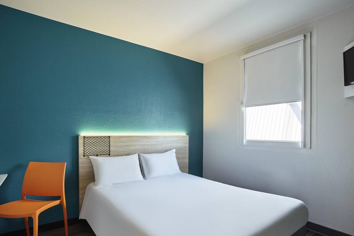 A hotel room with a bed, a chair, a window, and teal walls.