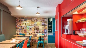Una cafetería con mesas de madera y sillas de colores, una barra roja con una máquina de café y una pared decorada con carteles de viaje.