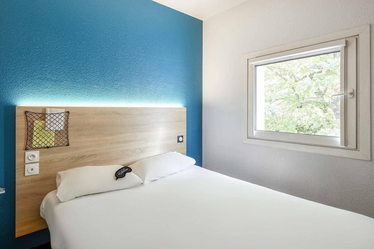 A hotel room with a wooden headboard, a white bed, a window with a view of greenery, and a power outlet.