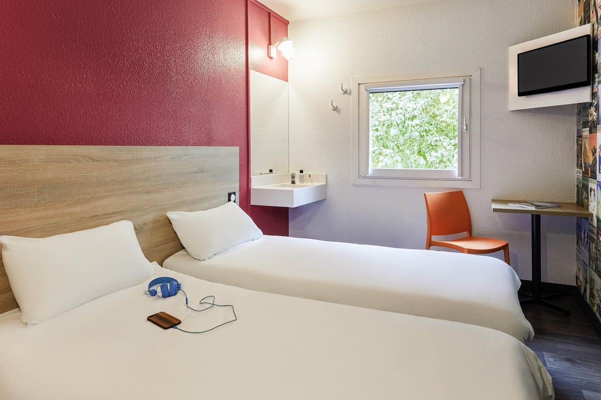 A hotel room with two beds, a desk, a chair, a TV, and a window with a view of greenery.