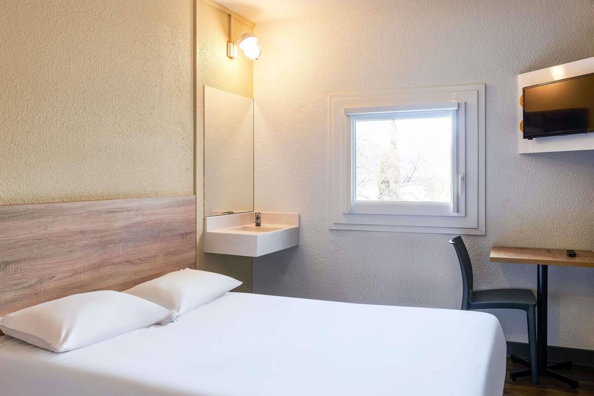 Een hotelkamer met een bed, nachtkastje, spiegel, wastafel en raam.