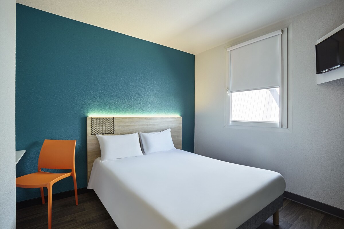 Una habitación de hotel con cama, una silla, un televisor y una ventana con persianas.