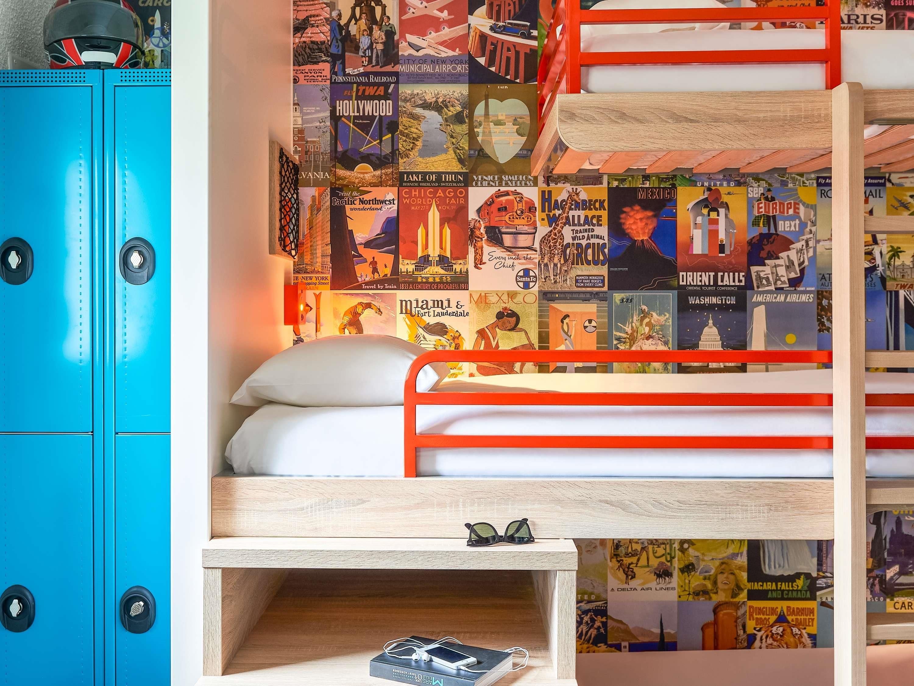 Una cama litera con cabecera de madera, una mesita de noche con un par de gafas de sol, y un armario azul con dos tiradores redondos.