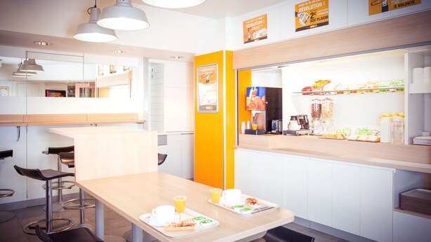 A modern café with a wooden table, bar stools, and a counter with food items. - hotelF1 Annemasse (Saint-Cergues)