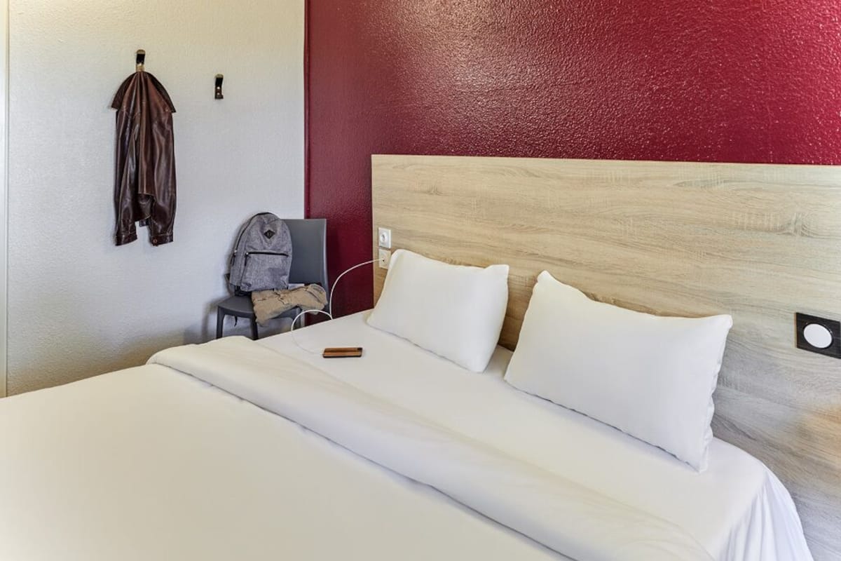 A hotel room with a large bed, a wooden headboard, a small desk with a chair, and a coat hanging on the wall.