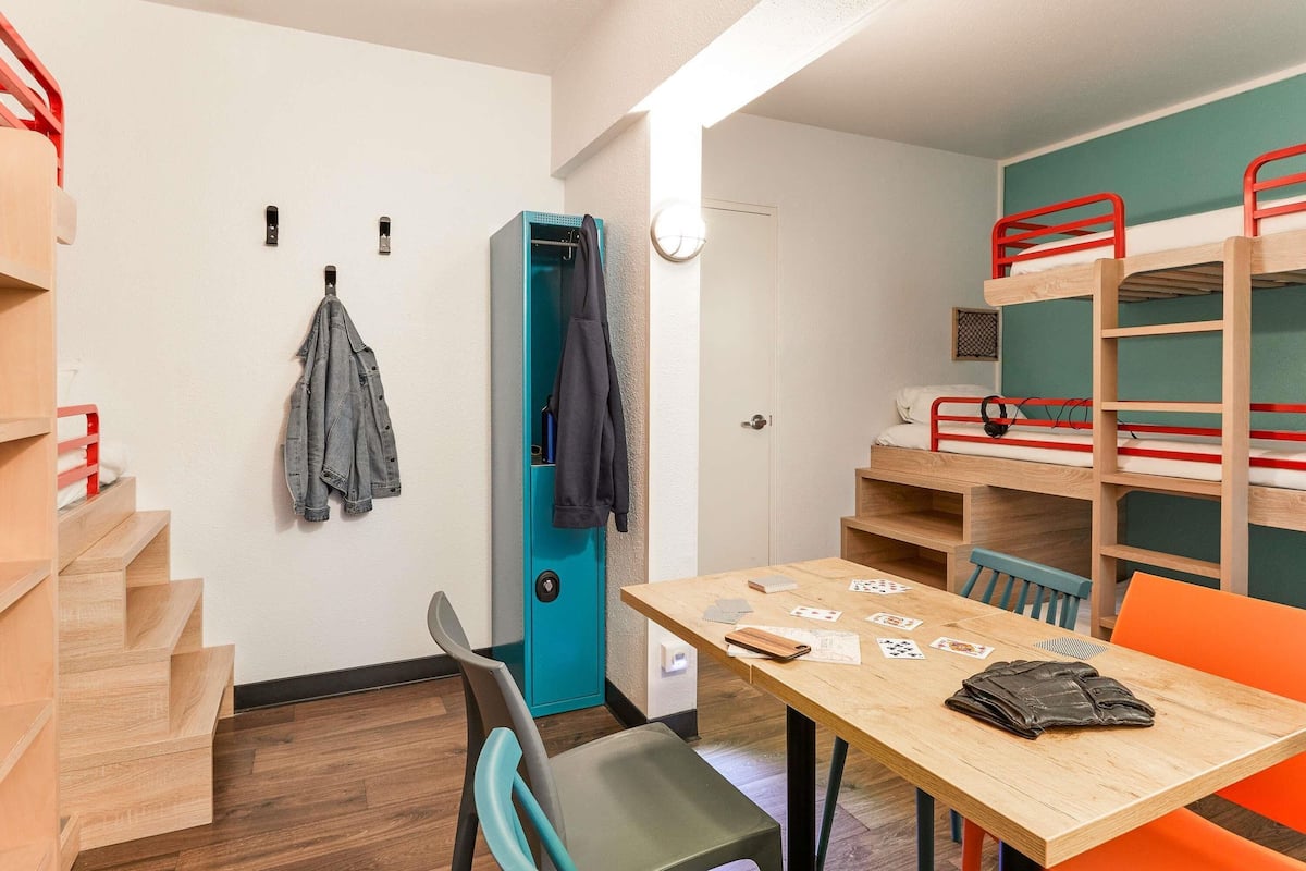 A dormitory room with bunk beds, a desk with cards, a chair, a coat rack, and a door.