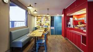 Un coin repas avec une longue table en bois, des chaises bleues et un banc. On y trouve des meubles bleus et un comptoir rouge avec une machine à café.