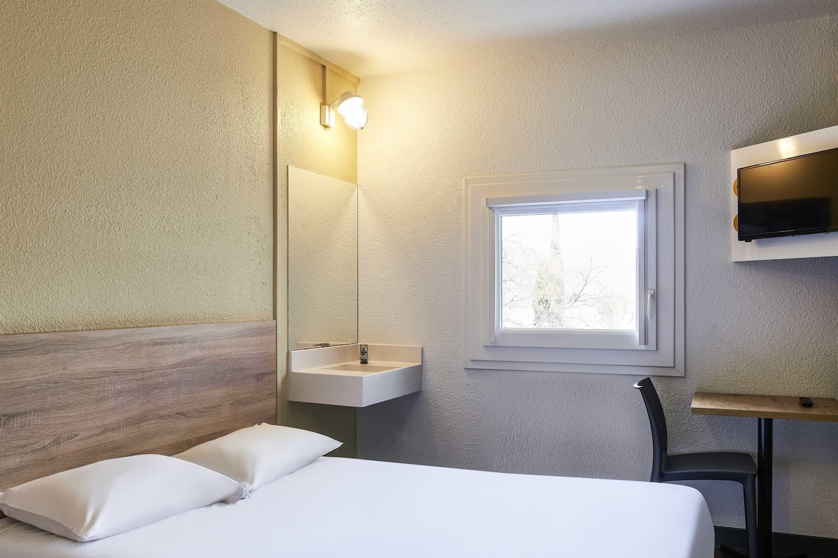 Habitación de hotel con cama, mesita de noche, espejo, lavabo y ventana.