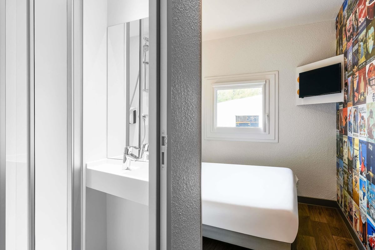 Un baño pequeño y moderno con ducha, lavabo e inodoro. Cuenta con una ventana que proporciona luz natural y un televisor instalado en la pared.