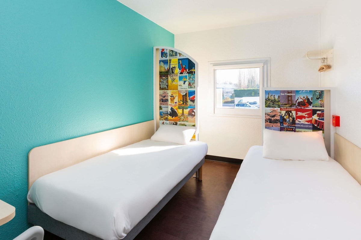 Une chambre d’hôtel avec deux lits simples, une petite table, une fenêtre donnant sur un paysage agréable et un mur décoré d’affiches de voyage.