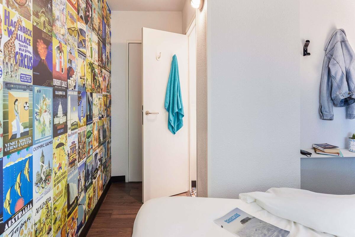 Une chambre d’hôtel avec un lit, un bureau doté d’une lampe, un mur orné d’affiches de voyage et une porte sur laquelle est accrochée une serviette.