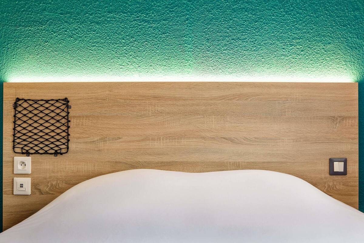 Une pièce bien rangée, avec une tête de lit en bois, un lit blanc, une étagère en treillis noir et une prise de courant au mur.