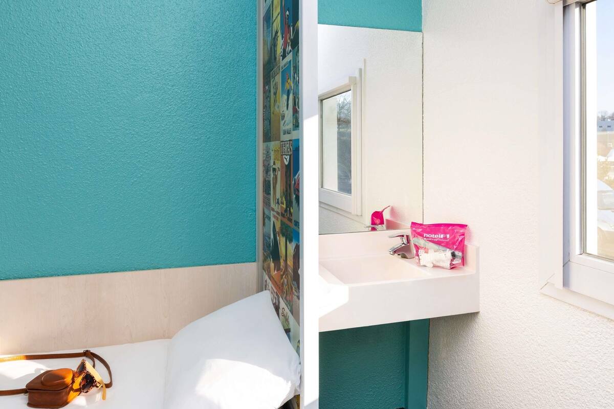 Une chambre d’hôtel aux murs de couleur turquoise, avec un lavabo blanc, un miroir et un lit avec un oreiller et un sac marron.