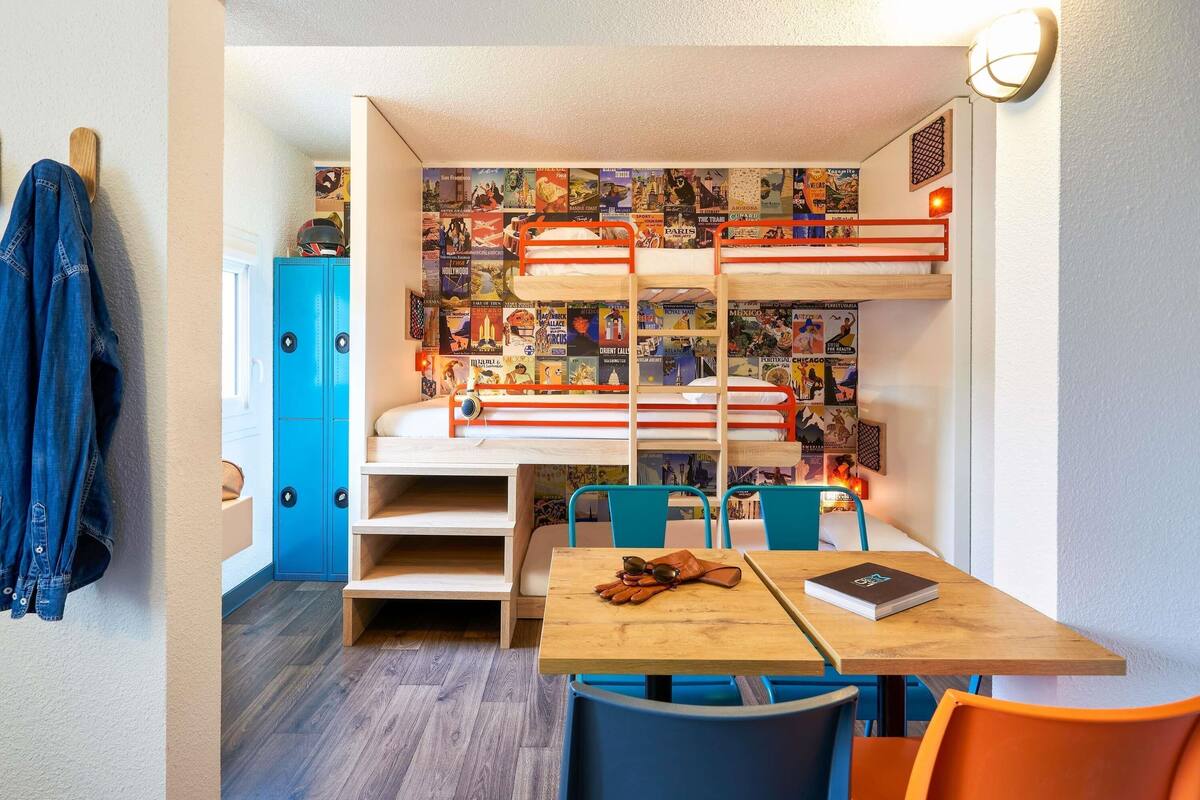 Une chambre compacte équipée de lits superposés, d’un bureau et de chaises, et dont un mur est décoré d’affiches sur le thème du voyage.