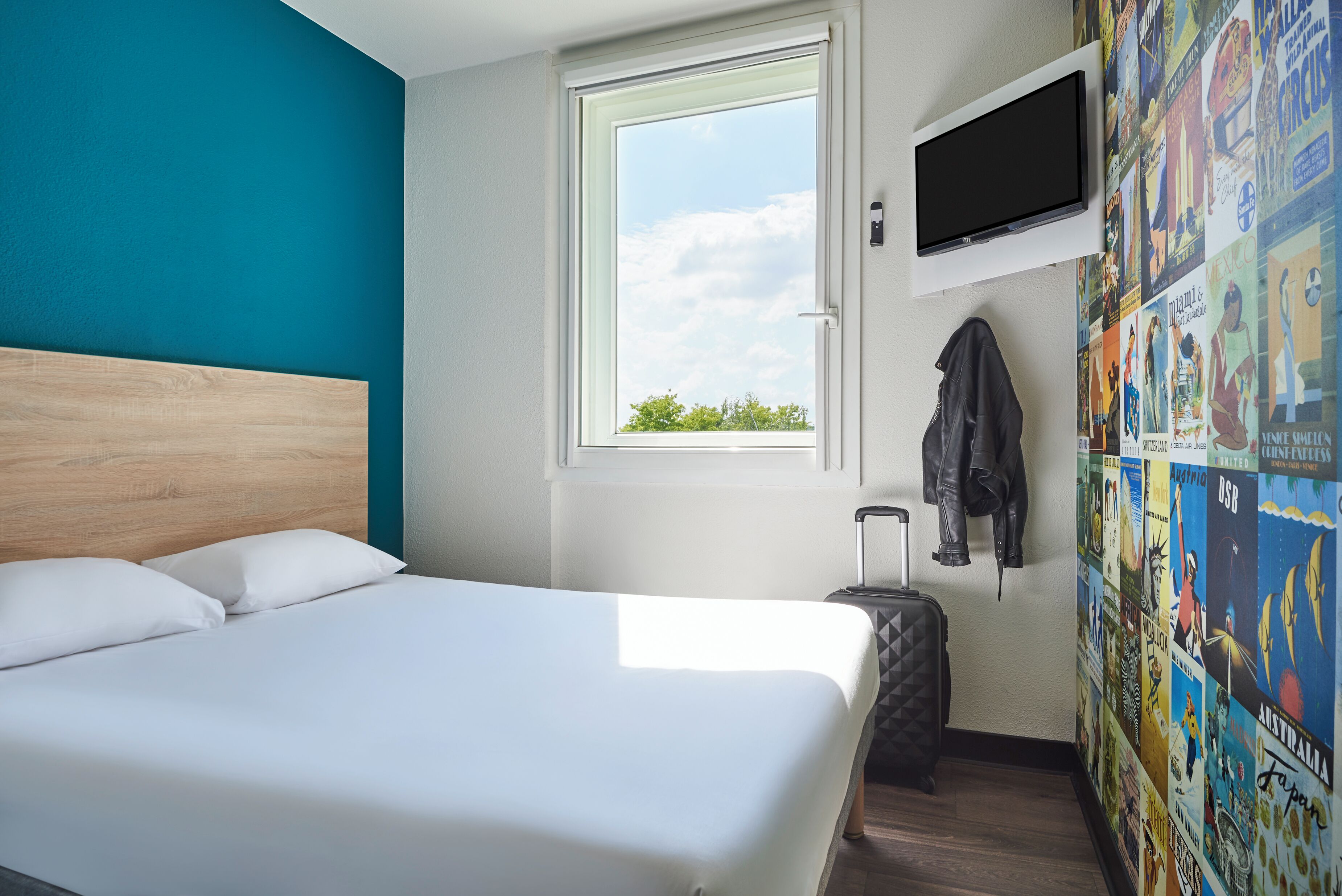 A hotel room with a bed, a window, a TV, and a wall with posters.