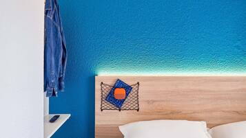 Una stanza con una testiera in legno, un letto con cuscini bianchi, una giacca di jeans appesa a una parete e un piccolo ripiano con un telecomando.