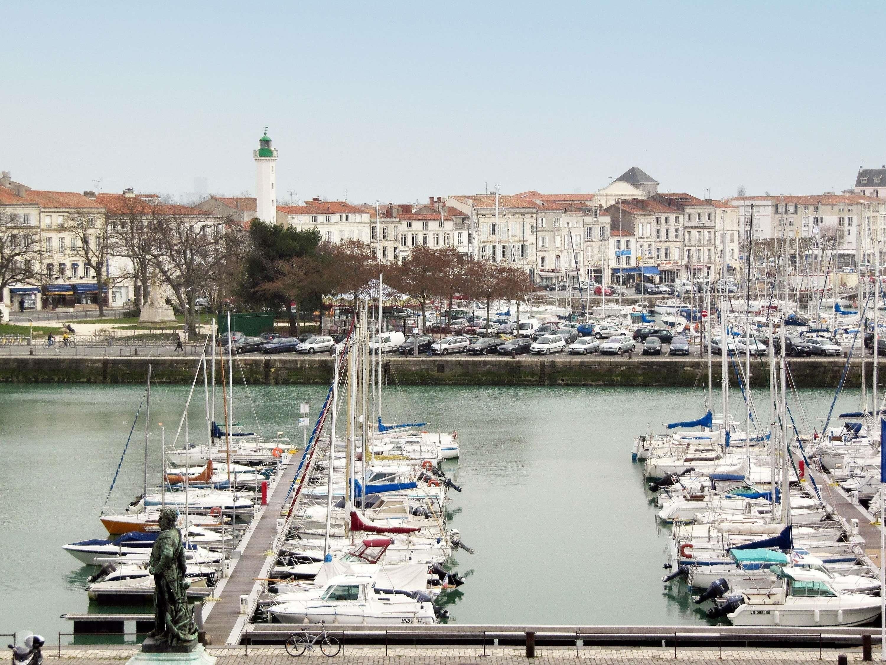 Un port de plaisance où sont amarrés de nombreux bateaux, une statue et un phare en arrière-plan.