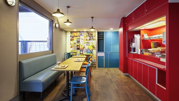 Un comedor con una mesa de madera larga, sillas azules y un banco. Hay armarios azules y una encimera roja con una máquina de café.