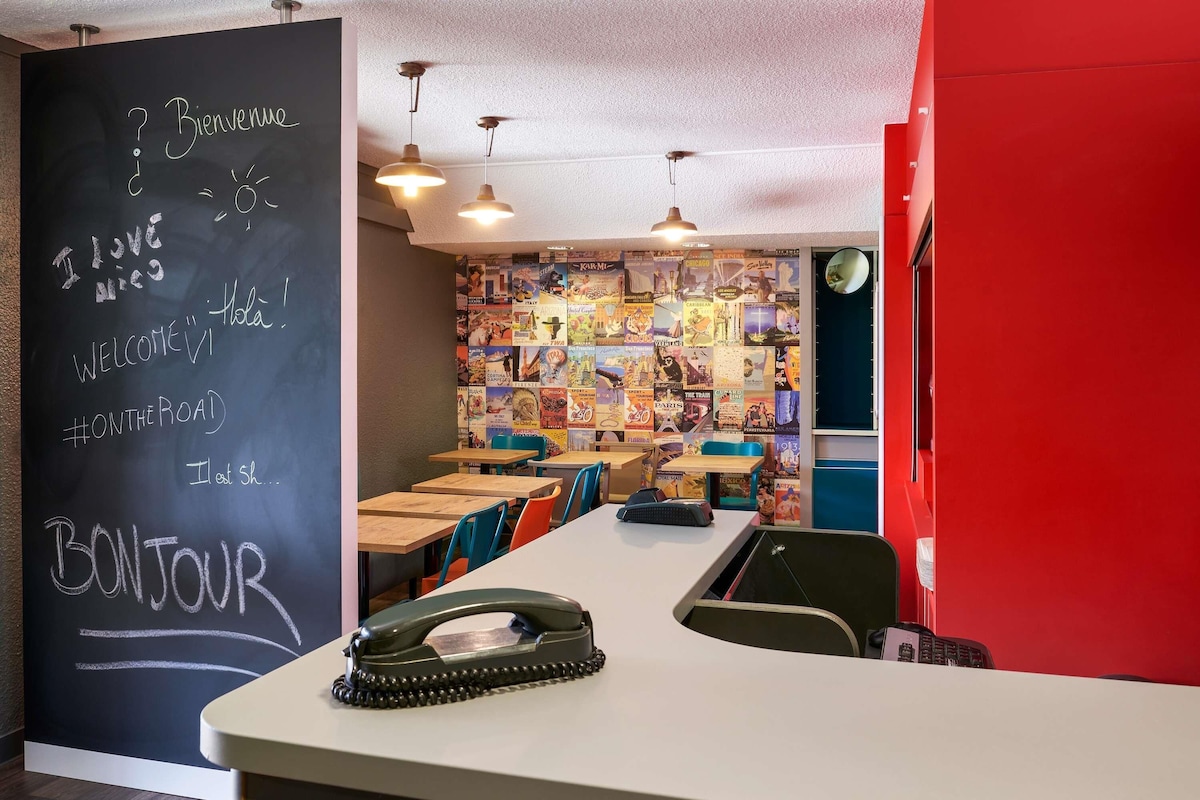 A modern cafe with a chalkboard wall displaying greetings and a hashtag, wooden tables and chairs, and a wall of colorful travel posters.