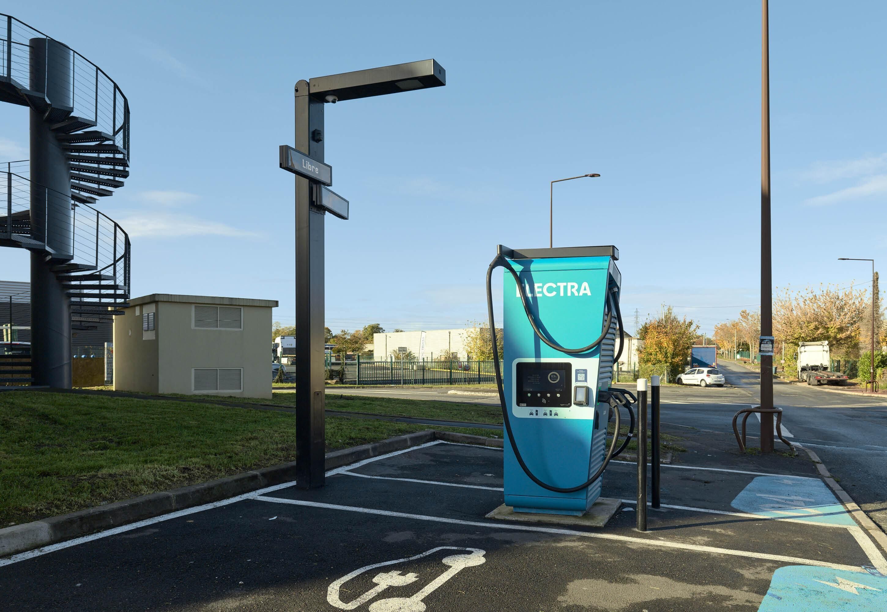 An electric vehicle charging station with a clear path for bicycles, a street lamp, and a building with a spiral staircase in the background.