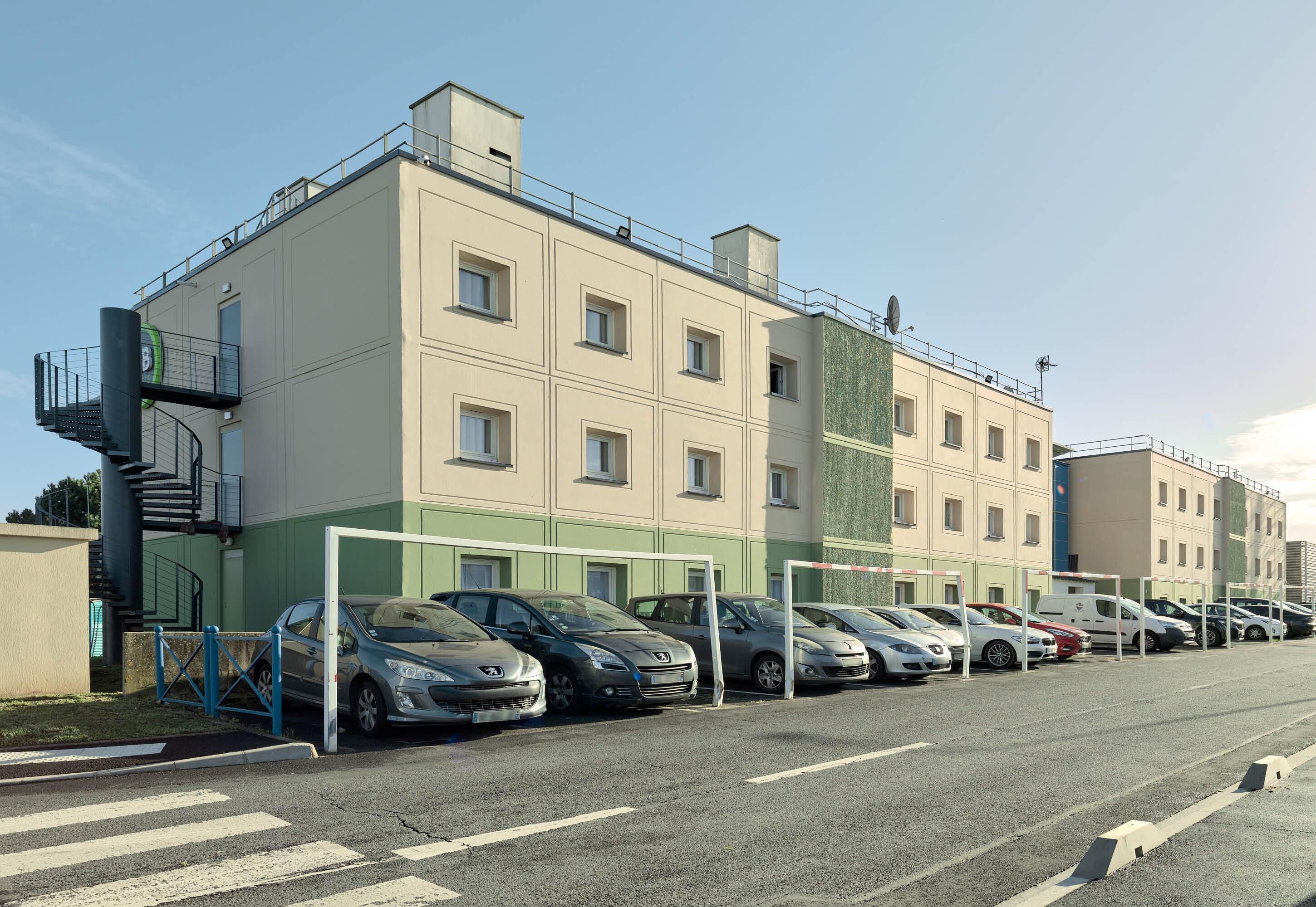 Un parking rempli de voitures, un bâtiment à la façade verdoyante et un ciel dégagé.
