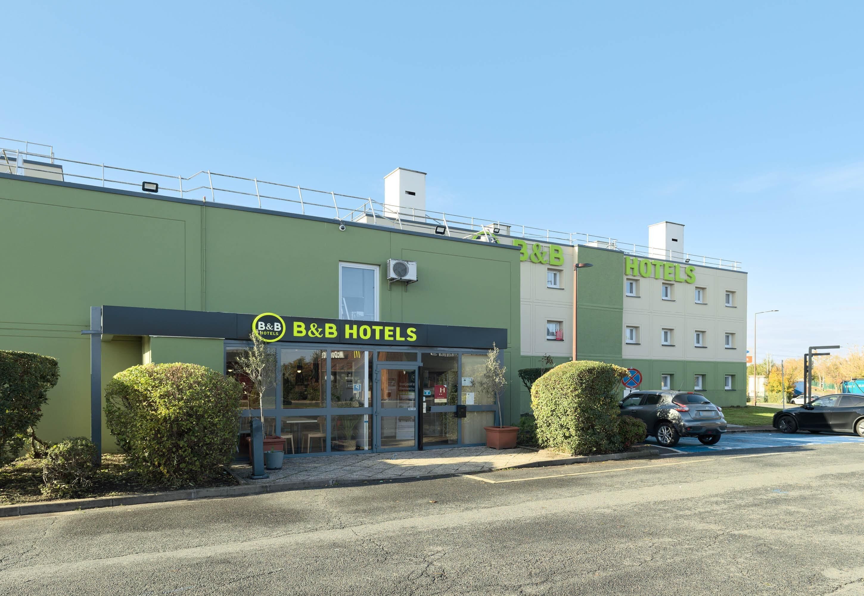 A B&B hotel with a green facade, a visible entrance, and a parking area with cars.